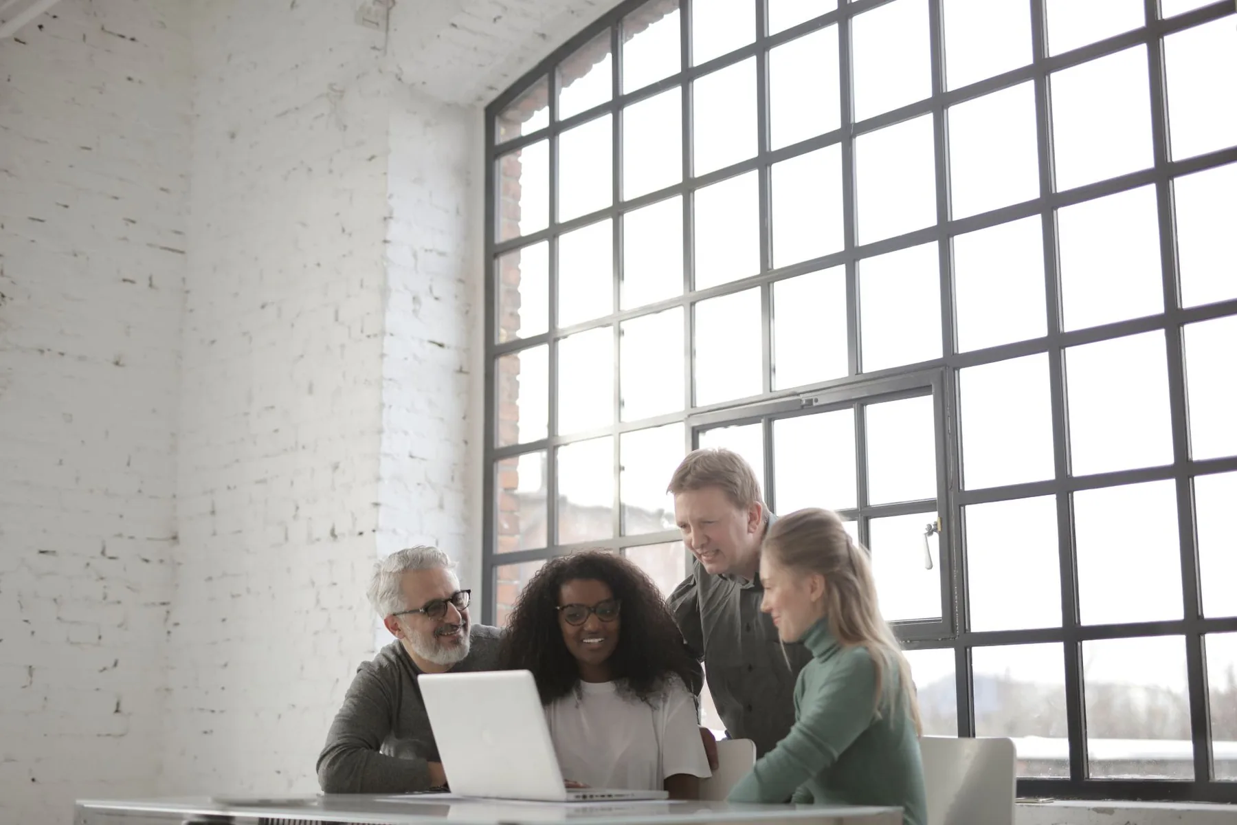Positive focused multiracial coworkers gathering together near table with laptop in workplace at industrial building during remote work on team against big window at daytime